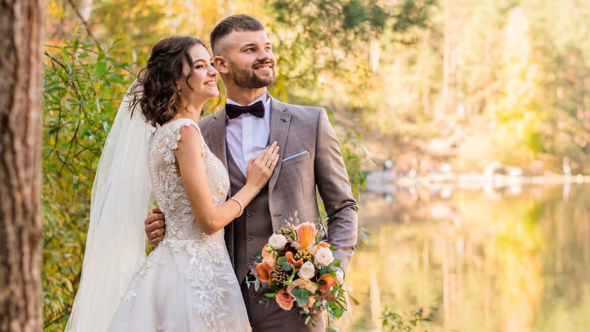 A smiling bride and groom holding each other beside a lake in a golden autumn forest